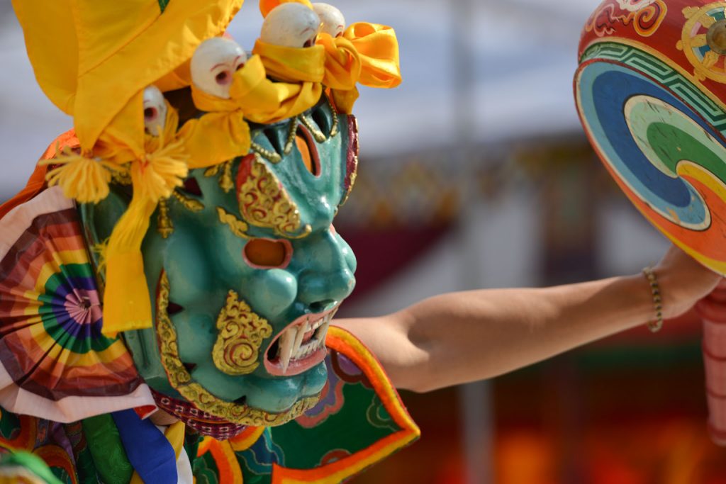 An elaborate ritual mask for the performanc e of a Cham Dance worn by a monk during a performance. The mask is green with rainbow coloured and gold details, and addorned with small sculptures of skulls.