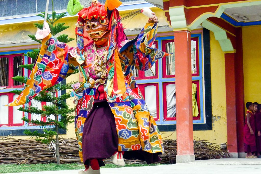 A monk swirl his robes of red, blue, green and gold as they perform a ritual Cham Dance against a mountain backdrop.